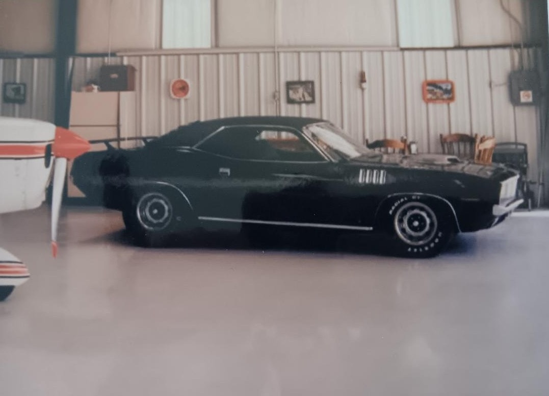 The black Plymouth 'Cuda parked inside Dan Powell's private hangar — his plane visible to the left, the 'Cuda gleaming on the polished concrete floor
