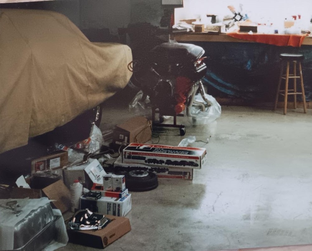 Dan Powell's Collinsville Oklahoma shop — 426 Hemi engine on a stand, parts boxes and components laid out across the floor, the 'Cuda covered in the background awaiting its heart