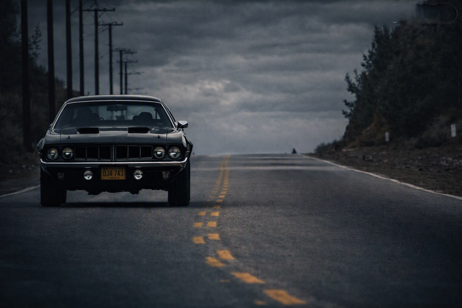 Black 1971 Plymouth Cuda coming straight down an empty two-lane road under a dark overcast sky — plate DJ4 743, quad headlights on, power lines running alongside, the road stretching to the horizon
